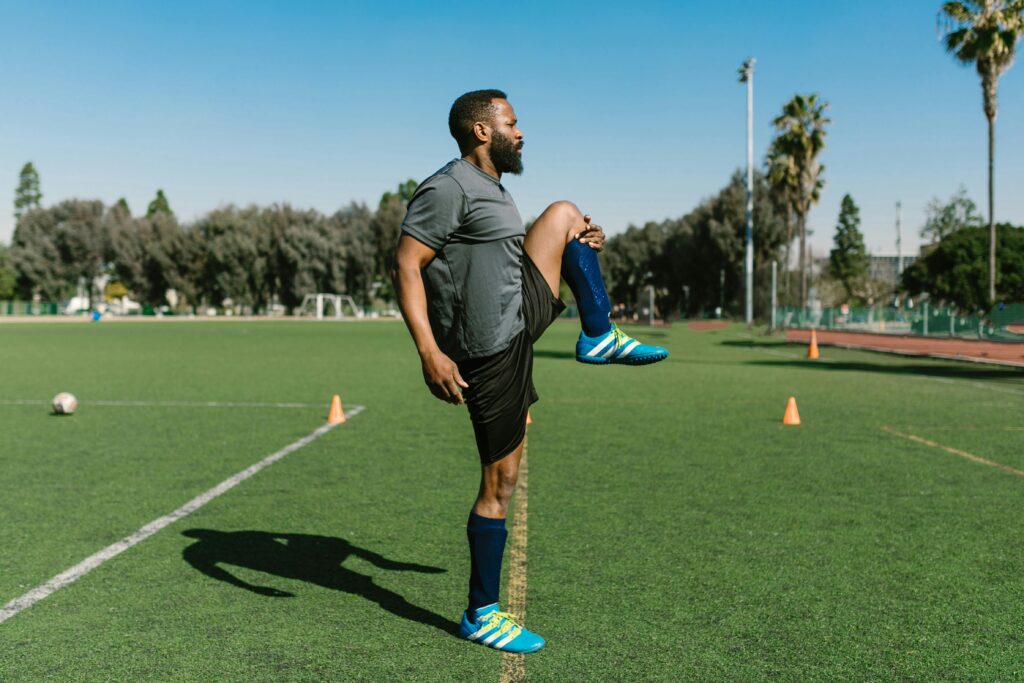 Black male athlete stretches on a green soccer field under a clear sky, preparing for training.